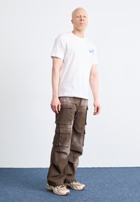White cotton T-shirt with a small blue logo, paired with brown cargo pants featuring multiple pockets. Shoes in beige with dark accents.