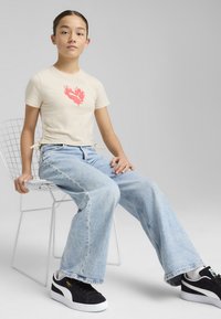 Camiseta beige claro con gráfico de llama roja, cordones a los lados, combinada con unos jeans de pierna ancha azul desgastados y zapatillas negras.