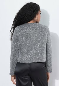 Vrouw met lang krullend haar en zilveren oorringen in de vorm van een ring, gekleed in een getextureerde zilveren lange mouw top en zwarte satijnen broek, met haar rug naar de camera gekeerd.