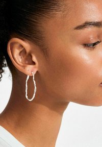 Orecchino a cerchio intrecciato in argento, caratterizzato da una forma organica e irregolare e da una finitura lucida e testurizzata, indossato su un design minimalista.