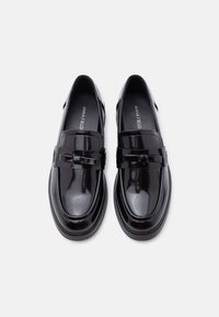 Ζευγάρι λαμπερών μαύρων παπουτσιών loafer από δερμάτινο γυαλιστερό πατέντα, με μικρό λουράκι και διακοσμητικό φιόγκο, που φαίνεται από πάνω σε λευκό φόντο.