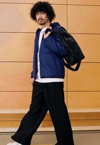 Marineblå quiltet jakke, hvit skjorte med striper, svarte vide bukser og svart ryggsekk. Bakgrunn med trepanel, lys gulvflate.