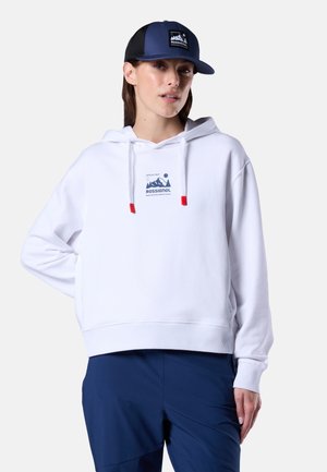 Kvinde iført en hvid Rossignol-hoodie med bjerlogo, marineblå bukser og en marineblå Rossignol-kasket med netbagside.