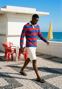 Homme portant une chemise à manches longues rayée rouge et bleue et un short blanc marchant sur un pavement carrelé près de chaises rouges et d’un parasol rayé jaune au bord de la mer.