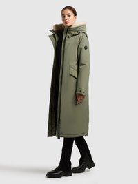 Lange, olijfgroene donsjas met een capuchon gevoerd met bont, zijzakken en een ritssluiting aan de voorkant. Gecombineerd met zwarte broek en stevige zwarte laarzen.