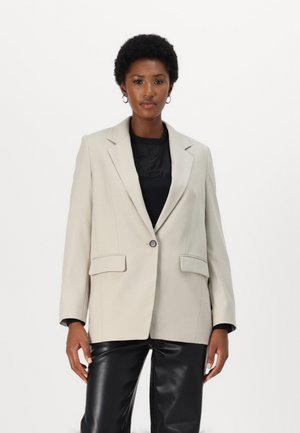 Beige taillierter Blazer mit Reverskragen, einreihigem Knopfverschluss, zwei vorderen Taschen und strukturierten Schultern, getragen über einem schwarzen Oberteil.