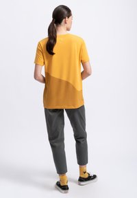 Korte mouwen gele t-shirt met een gebogen kleurblokontwerp, gecombineerd met grijze broek en gele geblokte sokken, gezien van achteren.