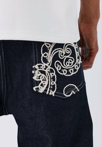 Spijkerbroek in donkerblauwe kleur met ingewikkelde witte borduursels op de achterzak, met draaiende patronen en contrasterend stiksel.