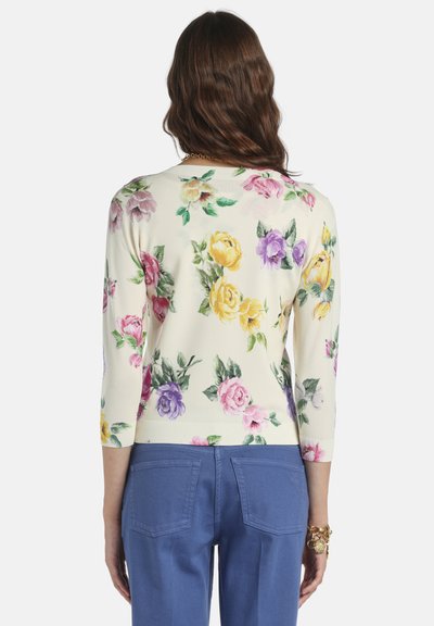 Virágmintás pulóver krémszínben, háromnegyedes ujjakkal. Sárga, rózsaszín és lila rózsákkal díszítve, bordázott szegéllyel. Kék nadrággal párosítva.