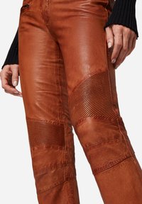 Bruine leren broek met ribbeltextuuraccenten op de knieën. Heeft een gladde afwerking en een op maat gemaakte pasvorm. Close-up van het stofdetail.