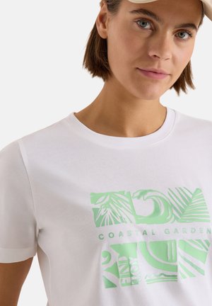 Femme portant un t-shirt blanc avec un motif abstrait vert clair de jardin côtier et une casquette beige, sur fond uni.
