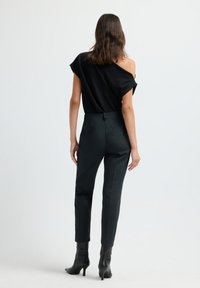 Femme portant un haut noir à épaules dénudées, un pantalon noir ajusté et des bottines noires à talons, se tenant contre un fond blanc, de dos face au spectateur.