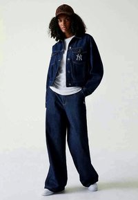 Τζάκετ από τζην με μπλε τελείωμα και λογότυπο NY, συνδυασμένο με φαρδιά μπλε τζιν. Λευκό μπλουζάκι από κάτω και καπέλο με καφέ σχέδιο. Λευκά παπούτσια.