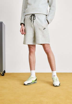 Jasnoszara bawełniana bluza i dopasowane szorty z czarnym logo Nike. Białe i zielone sneakersy, białe skarpetki do kostek oraz żółte tło z dywanem.