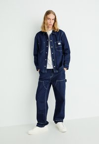 Giacca in denim blu scuro con due tasche frontali e cuciture bianche, abbinata a pantaloni coordinati e sneaker bianche. Texture liscia in tutto.