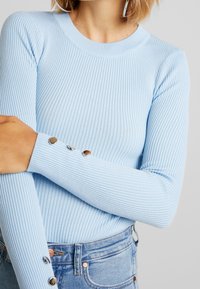 Top de manga larga de color azul claro con relieve, con un escote redondo y detalles de botones en los puños, combinado con pantalones cortos de mezclilla de tiro alto.