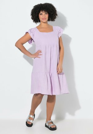 Mujer con cabello rizado, vestida con un vestido lila hasta la rodilla y sandalias plateadas, de pie con una mano en la cadera frente a un fondo blanco.