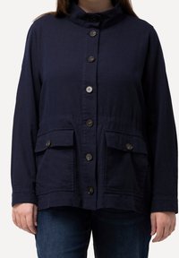 Kvinna som bär en marinblå knäppbar jacka med ståkrage och två fickor med klaff, kombinerad med mörkblå jeans.