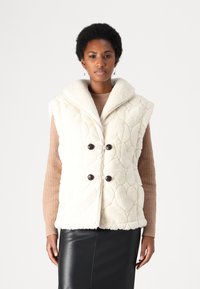Faux fur-väst i off-white med ett kviltat hjärtmönster, bred krage och två mörka knappar. Lagd över en ribbad brun topp.