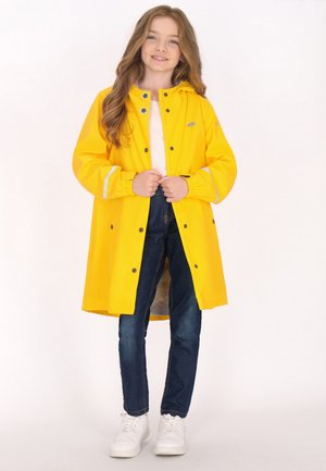 Gelbe wasserdichte Regenjacke mit einem Reißverschluss vorne, Druckknöpfen und reflektierenden Akzenten, kombiniert mit blauen Jeans und weißen Sneakers.