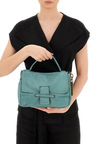 Borsa in pelle turchese con manico superiore, forma rettangolare, chiusura con pattina frontale e dettaglio decorativo sulla tracolla. Presenta un'etichetta con accentuazione dorata.