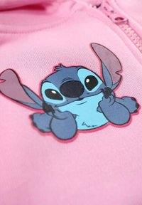 Sweat à capuche rose clair avec un personnage de dessin animé cousu, aux accents bleus et noirs, grands yeux et bras expressifs.