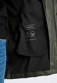 Interior de chaqueta impermeable verde con forro negro, que presenta etiquetas que indican propiedades impermeables y a prueba de viento. Tejido texturizado y acabado suave.