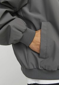 Gray bomber jacket featuring a smooth, matte finish, elastic cuff, and side pockets, with gathered hem for added shape and structure.