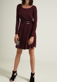 Burgundowa sukienka z długim rękawem, z paskiem w talii, zwiewnym materiałem i okrągłym dekoltem, zestawiona z czarnymi szpilkami na kostce i okrągłą torebką.