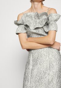 Femme croisant les bras, portant une robe argentée décolletée avec un col à volants et des motifs texturés ressemblant à des fleurs.