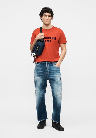 Νέος άνδρας που φοράει κόκκινο κοντομάνικο με γραφικά, ξεφτισμένα μπλε τζιν, μαύρα αθλητικά παπούτσια και μια μπλε τσάντα χιαστί, στέκεται με το ένα χέρι στην τσέπη.