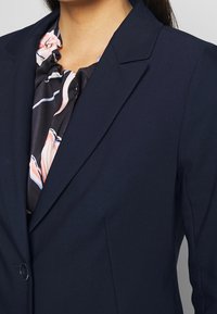Námořnický blejzr se strukturovanými rameny, klopami a zapínáním na knoflík. Pod ním je černá blouse s květinovým vzorem. Hladký povrch.