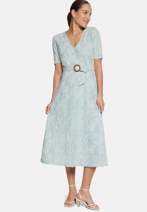 Žena v svetlomodrých midi šatách s bielym kvetinovým vzorom, krátkymi rukávmi, výstrihom do V, opaskom s okrúhlou prackou a béžovými sandálmi na podpätku.