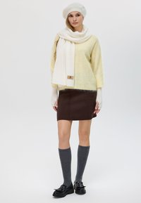 Maglione giallo chiaro, sciarpa bianca, gonna marrone scuro, calze grigie fino al ginocchio e scarpe nere. Morbidi tessuti e vestibilità rilassata in tutto.