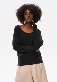 Schwarzes Langarmoberteil mit weitem Ausschnitt und Paillettenakzenten an der Seite, kombiniert mit einem fließenden beigen Rock. Glatte, weiche Textur.