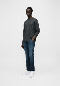 Γκρι μακρυμάνικο μπλουζάκι με λογότυπο, σκούρο μπλε τζιν και λευκά αθλητικά παπούτσια. Casual εφαρμογή και βαμβακερό υλικό, στέκεται μπροστά σε απλό φόντο.