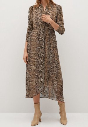 Frau trägt ein langärmliges, midi-langes Kleid mit Schlangenledermuster und beigefarbene Stiefeletten, steht vor einem schlichten hellen Hintergrund.