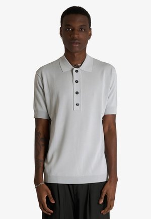 Giovane uomo che indossa una polo grigio chiaro a maniche corte con quattro bottoni neri e pantaloni neri, in piedi davanti a uno sfondo semplice.