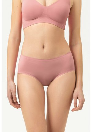 Vrouw draagt naadloze lichtroze beha en bijpassende hoog uitgesneden slip, staand met ontspannen armen langs het lichaam tegen een witte achtergrond.