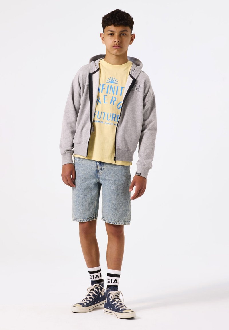 Sweat à capuche gris zippé, t-shirt graphique jaune, shorts en denim bleu, baskets marines et chaussettes à logo noir et blanc. Style streetwear décontracté.