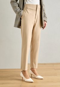 Béžové kalhoty na míru se slim fit, vysokým pasem a předními sklady, doplněné o bílé lodičky s špičatou špičkou a kostkovaným blejzrem.