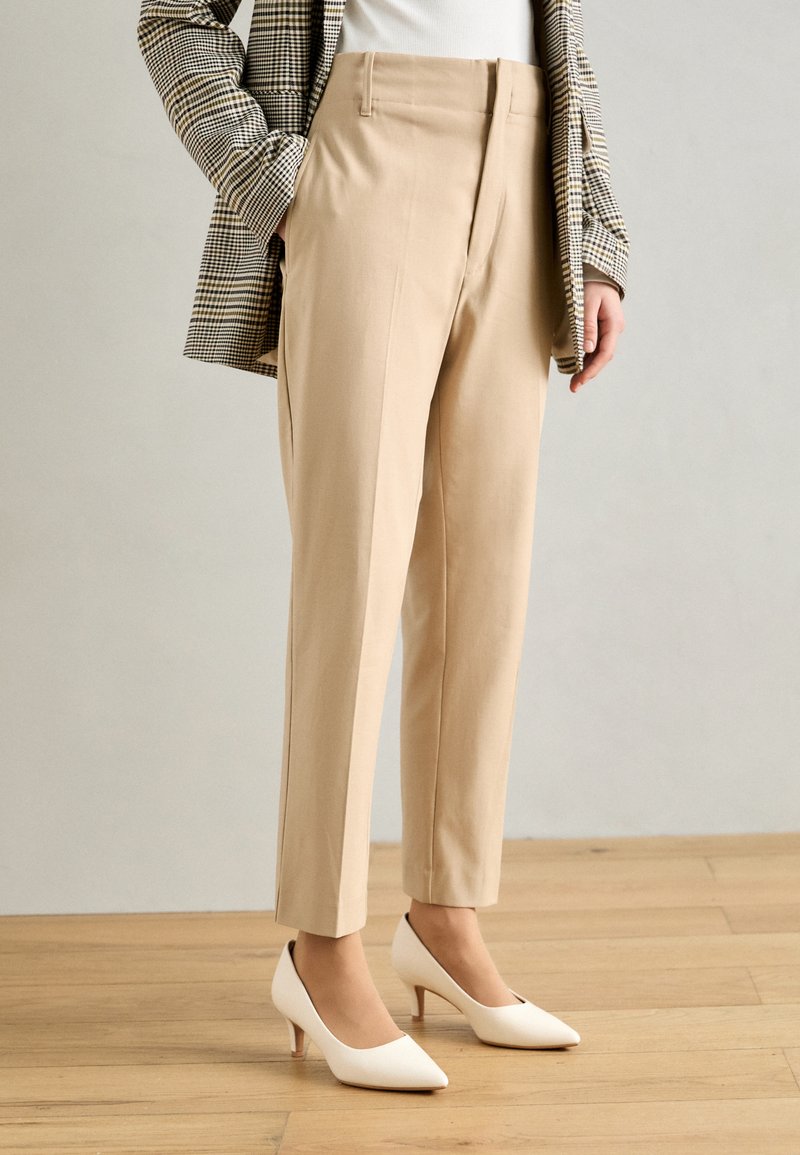 Béžové kalhoty na míru se slim fit, vysokým pasem a předními sklady, doplněné o bílé lodičky s špičatou špičkou a kostkovaným blejzrem.