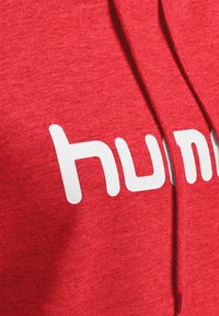 Haut à capuche rouge avec une texture douce, comportant un texte logo blanc bien visible. Le design comprend une capuche et des cordons de serrage, mettant l'accent sur le style décontracté.