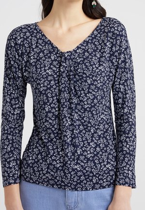 Vrouw draagt een marineblauwe top met lange mouwen met een wit bloemenpatroon en geplooide details in het midden vooraan, gecombineerd met lichtblauwe spijkerbroek.