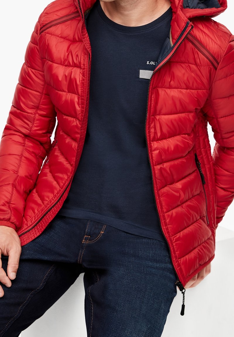 Veste doudoune rouge avec texture matelassée, accents gris et fermeture éclair à l'avant. Portée sur un t-shirt marine et un jean bleu foncé avec des coutures orange.