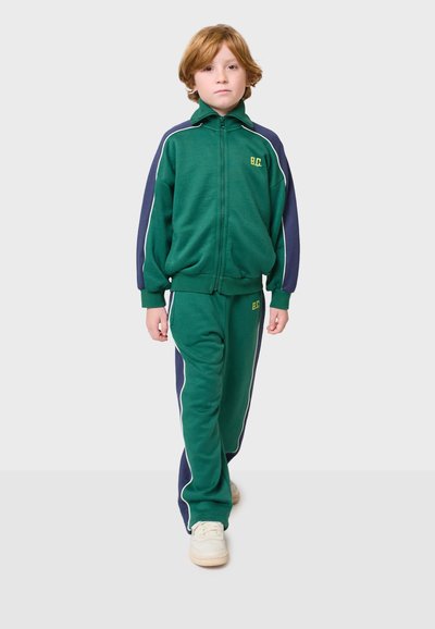 Zeleni trenirka od mekanog materijala, koja uključuje jaknu na zatvaranje s patentnim zatvaračem s mornarskim naglascima i odgovarajuće hlače. Sadrži bijele bočne pruge i logo.