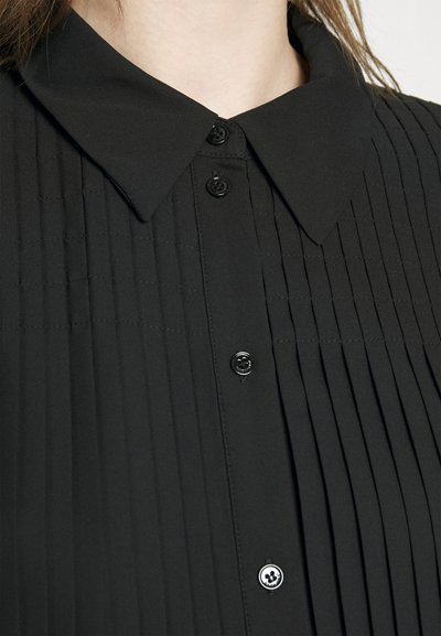 Bluza neagră plisată cu guler, cu butoane negre și o textură netedă. Plisaje verticale notabile pe întreaga țesătură.