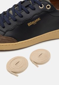 Sneaker in pelle blu navy con dettagli in suede marrone, logo dorato e lacci in tessuto. Due lacci rotondi beige mostrati accanto alla scarpa.