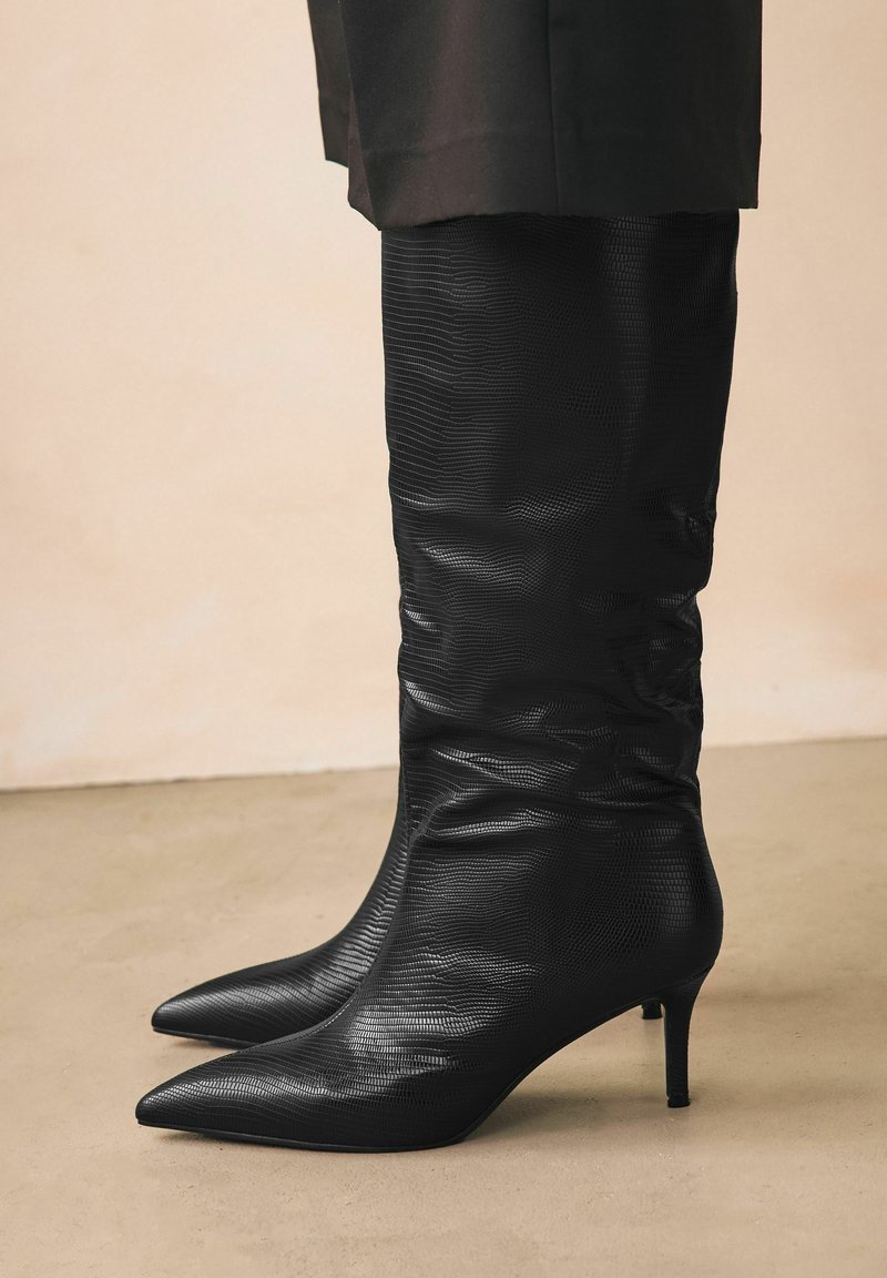 Stivali neri al ginocchio realizzati in materiale testurizzato con un motivo sottile. Caratterizzati da una punta affusolata e un tacco sottile per una silhouette elegante.