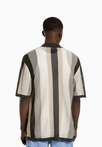 Camisa de manga corta a rayas en tejido de malla negro, crema y gris. Cuenta con un cuello negro y un corte holgado con costuras visibles.